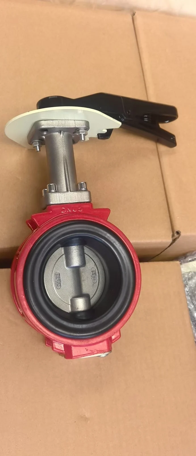 A red-bodied butterfly valve with a black hand lever, photographed on cardboard packaging